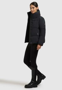 Donna in piedi con giacca imbottita nera, leggings neri e stivali neri alti fino al ginocchio, su uno sfondo chiaro e uniforme.