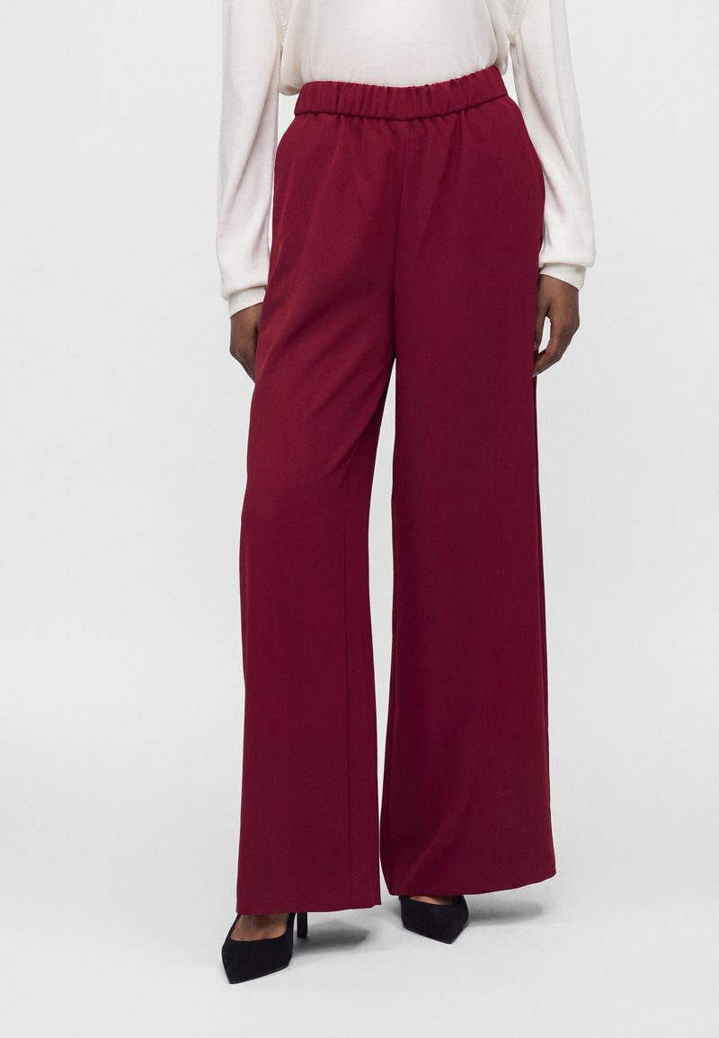 Personne portant un pantalon bordeaux taille haute, jambes larges, taille élastique, et des talons noirs à bout pointu sur un fond uni.