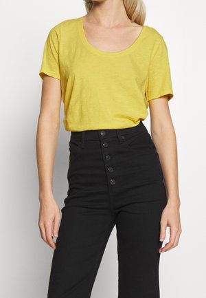 Żółta koszulka z krótkim rękawem i dekoltem typu woda, noszona z wysokim stanem czarnych spodni z guzikami i gładką fakturą.