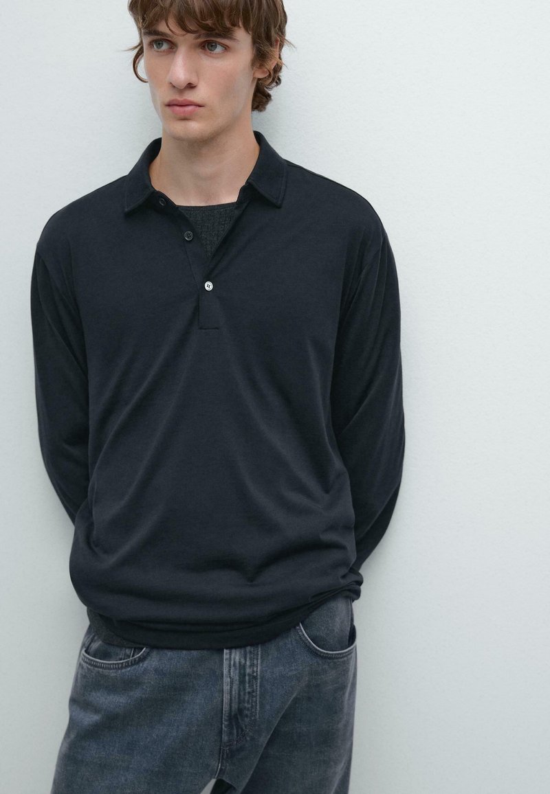 Giovane uomo con capelli castano chiaro che indossa una polo a maniche lunghe scura sopra una maglietta nera e jeans blu sbiaditi, in piedi contro un muro bianco.