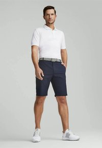 Bela polo majica, tamnoplave kratke hlače i bijele tenisice. Pojas ima teksturirani srebrni dizajn. Model stoji s jednom rukom u džepu.