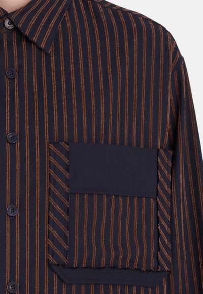 Chemise boutonnée bleu marine avec des rayures verticales marron, présentant une grande poche plaquée avec des rayures diagonales et un design de col structuré.