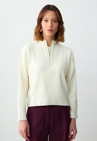 Pull en maille côtelée couleur crème avec un zip quart à l'avant, des manches longues et une coupe décontractée. Stylé avec un pantalon bordeaux profond.
