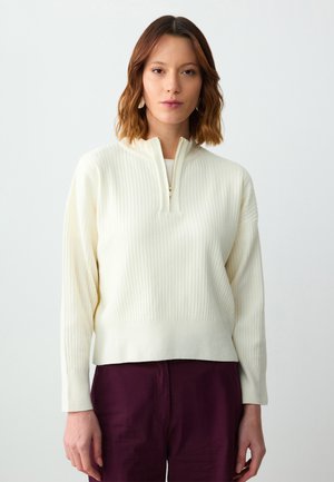 Crème kleurige geribbelde pullover met een kwart rits aan de voorkant, lange mouwen en een relaxte pasvorm. Gestyled met diep bordeauxrode broek.