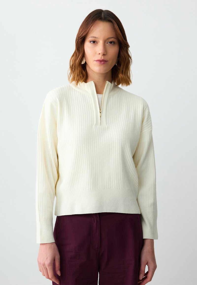 Pull en maille côtelée couleur crème avec un zip quart à l'avant, des manches longues et une coupe décontractée. Stylé avec un pantalon bordeaux profond.