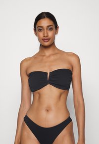 Črn bandeau bikini zgornji del z teksturiranim materialom in središnjo gumbovo podrobnostjo, v kombinaciji z nizkimi črnimi bikini hlačkami.