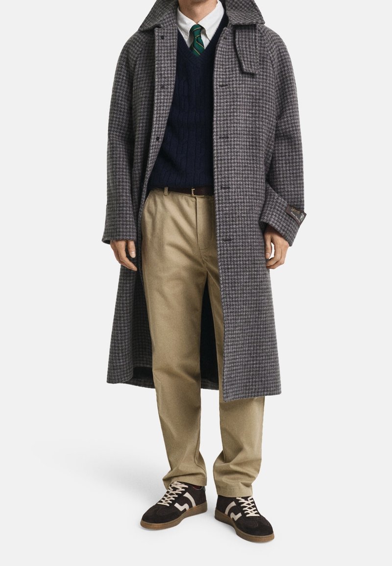 Homme portant un long manteau gris à motif pied-de-poule, un pull bleu marine, une chemise blanche avec une cravate rayée verte, un pantalon beige et des baskets noires à lacets blancs.