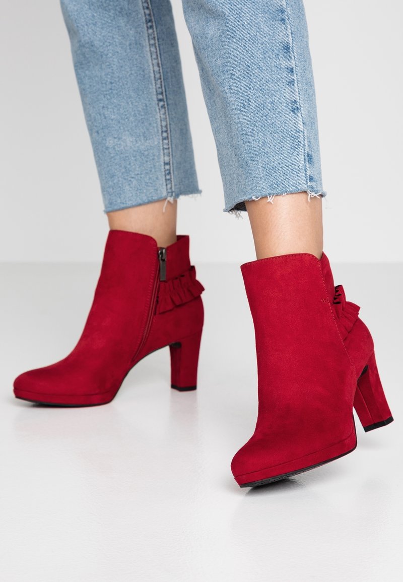 Bottines en suède rouge avec talons carrés et fermetures éclair latérales, portées avec un jean court bleu clair sur une surface blanche.