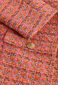 Geweven stof in oranje en roze tinten met een patroon; heeft een rond houten knoopaccent.