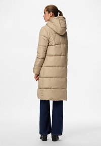 Beige dunjakke med hette, quiltet design, lange ermer og kne-lengde silhuett. Kombinert med mørkeblå vide jeans og ankelstøvler.