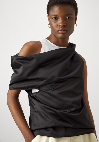 Top in satin nero con design drappeggiato, dotato di scollo a spalla scoperta e particolare con ritaglio visibile. Il modello indossa un sottogonna bianca.
