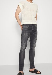 Suéter blanco de manga corta tejido; jeans ajustados gris oscuro con detalles desgastados; sandalias negras tipo slide; atuendo casual.