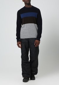 Pull tricoté avec des rayures horizontales bleu marine, bleu, marron et gris, avec des poignets et un ourlet côtelés. Associé à un pantalon noir ample et des bottes noires.