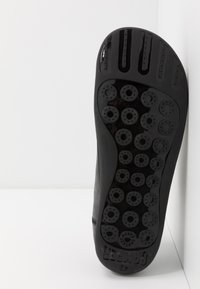 Suela de zapatilla negra con patrones circulares de agarre y orificios de ventilación. La parte superior lisa se transforma en una suela de goma texturizada con detalle del logo.