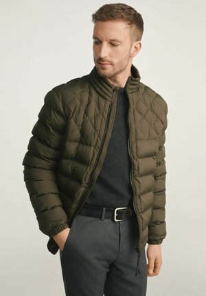 Pikowana oliwkowa kurtka puchowa z kołnierzem stojącym i zamkiem błyskawicznym, noszona na ciemnym swetrze. Sparowane z szarymi spodniami i czarnym paskiem.
