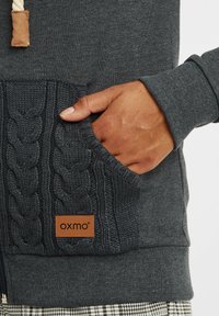 Mörkgrå hoodie med en flätad stickad ficka som har en läderlogotyp. Tyget har en jämn textur; fickan ger visuell kontrast.