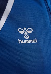 Primo piano di tessuto sportivo blu con logo bianco hummel che presenta un'ape stilizzata e testo nella zona del petto.
