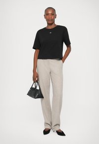 Cămașă neagră scurtă, cu un logo mic, pantaloni bej cu textură, largi, și o geantă din piele neagră. Modelul stă cu mâinile pe șolduri.