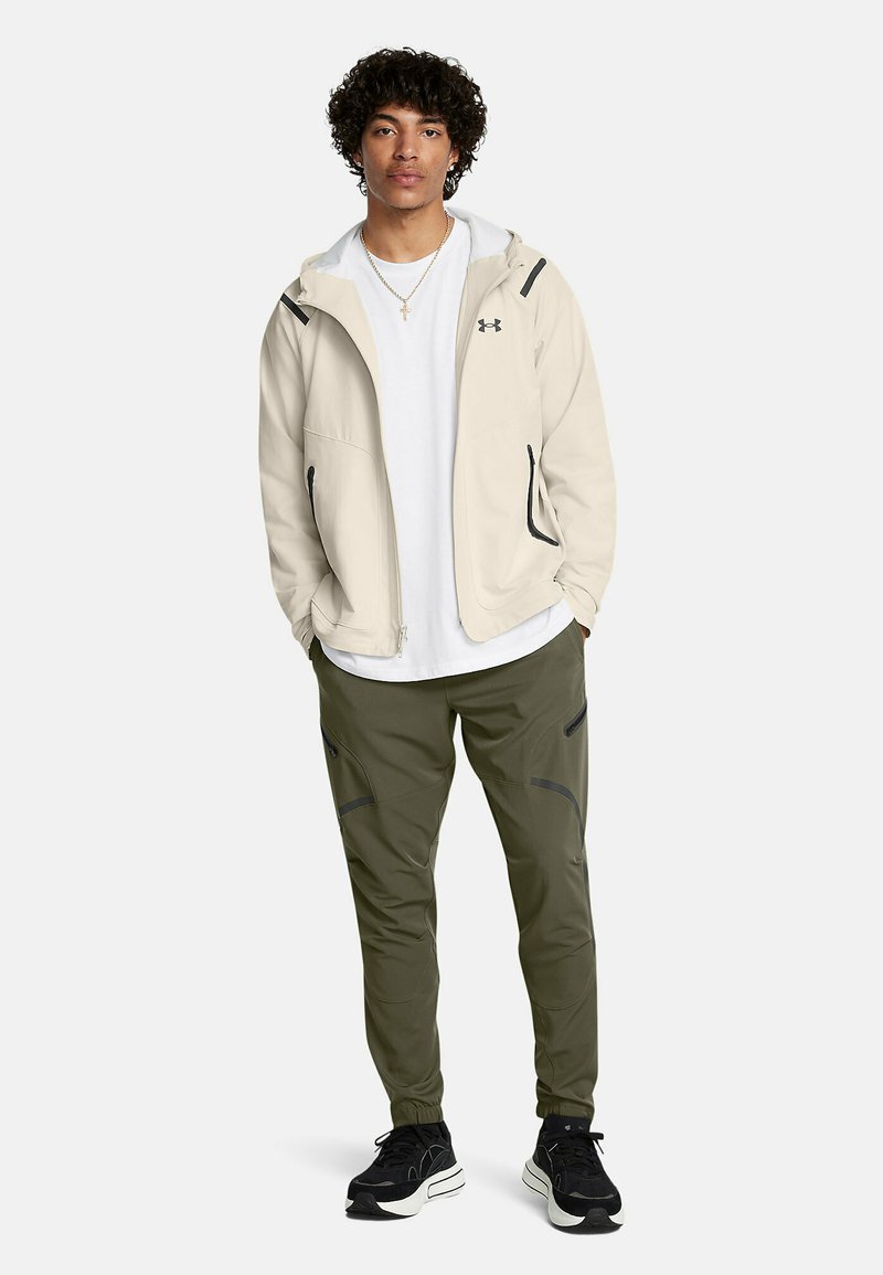 Veste beige zippée avec capuche, poches avant et accents noirs, associée à un pantalon vert olive avec des coutures courbes et des zippers noirs.