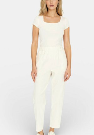 Kobieta ubrana w dopasowany biały krótki rękaw, białe spodnie z wysokim stanem oraz białe wsuwane buty, stojąca na tle jednolitego tła.