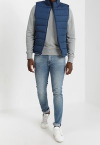 Marineblauer gepolsterter Weste über einem grauen Sweatshirt, kombiniert mit hellblauen Jeans und weißen Sneakern. Einfaches Design mit kontrastierenden Farben.
