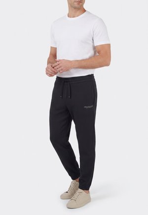 Mann trägt ein weißes kurzärmeliges T-Shirt, schwarze Kordelhosen mit Armani Exchange-Logo und beige Sneaker, steht vor einem einfarbigen Hintergrund.