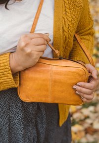 Person som bär en senapsfärgad stickad kofta och vit skjorta, håller och öppnar en liten tan-färgad lädercrossbody-väska utomhus.