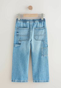 Pantalon cargo en denim bleu clair avec taille élastique, deux poches plaquées arrière et poches utilitaires latérales suspendu à un cintre en bois.