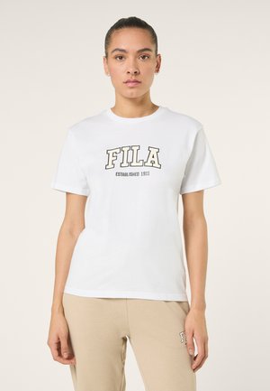 Kobieta z upiętymi włosami stoi przodem, ubrana w białą koszulkę FILA i beżowe spodnie z logo FILA.