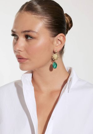 Goldfarbene Ohrringe mit einem Sonnendesign und grünen Edelsteintropfen. Das Modell trägt ein weißes Karierthemd. Glatte Texturen, kontrastierende Farben.