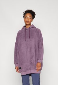 Overdimensjonert hettegenser i myk lilla fleece. Har ribbestrikkede mansjetter og en liten svart etikettdetalj ved sømmen. Jevn utvendig tekstur.