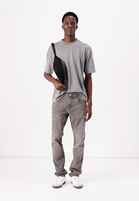 Tricou gri din bumbac, blugi gri și adidași albi. Modelul ține o geantă de umăr neagră cu o curea, stând în fața unui fundal simplu.