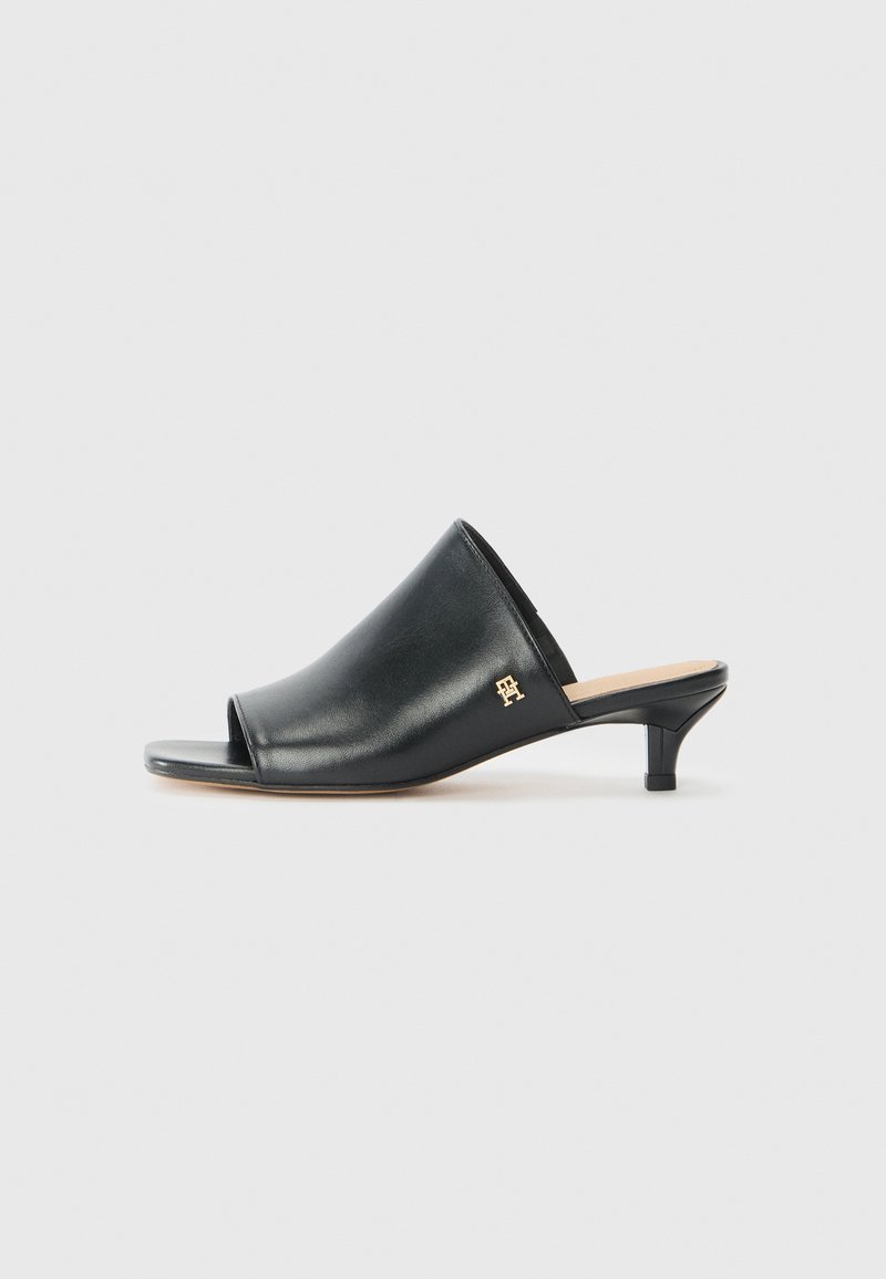 Mules de cuero negro de punta abierta con un tacón bajo. Presentan una superficie lisa y un acento del logo dorado en el costado.