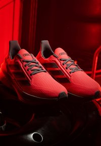 Paire de chaussures de sport rouges vives avec des rayures noires et des lacets gris, présentées sur un fond rouge foncé.
