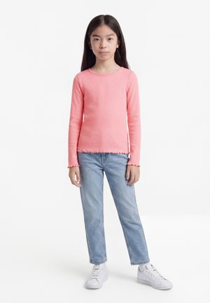 Κορίτσι με μακριά σκούρα μαλλιά στέκεται φορώντας ροζ μπλούζα με μακριά μανίκια, ανοιχτόχρωμα μπλε τζιν και λευκά αθλητικά παπούτσια, μπροστά σε λευκό απλό φόντο.