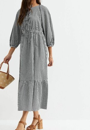 Žena v černo-bílých kostkovaných midi šatech s nafouklými rukávy, držící pletenou kabelku a nosící hnědé sandály na podpatku.