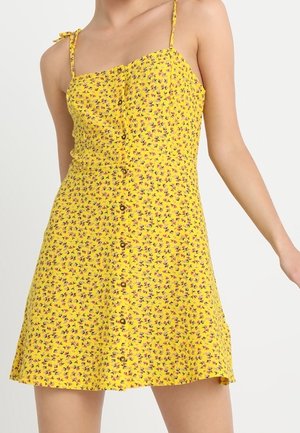 Donna che indossa un vestito giallo senza maniche con una piccola stampa floreale e bottoni sul davanti, in piedi contro uno sfondo chiaro e uniforme.