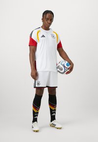 Λευκή φανέλα ποδοσφαίρου με κόκκινες και κίτρινες λεπτομέρειες, μαύρα σορτς, μαύρες κάλτσες μέχρι το γόνατο με σχέδιο, και λευκά παπούτσια. Ο παίκτης κρατάει μία μπάλα.