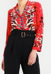 Red floral button-up blouse with long sleeves and a collar, featuring blue and white floral accents. Worn with high-waisted black trousers.