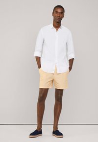 Chemise blanche en lin à manches longues, shorts rayés jaunes et chaussures à enfiler bleu marine. La tenue présente un design décontracté et léger pour l'été.