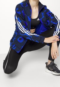 Chaqueta deportiva azul con estampado y rayas blancas, llevada sobre un sujetador deportivo negro y combinada con leggings negros y zapatillas oscuras.