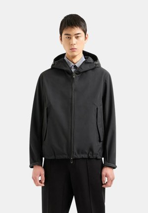 Giacca nera con zip e cappuccio, dotata di tasche laterali e coulisse regolabili. Texture liscia con un design leggero, adatta per essere indossata a strati.