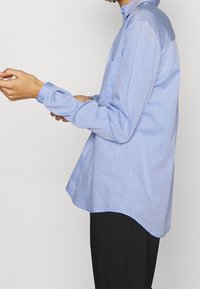 Blue long-sleeve shirt with a collar, featuring a light polka dot pattern. Made of smooth fabric, it has a relaxed fit and a breast pocket.