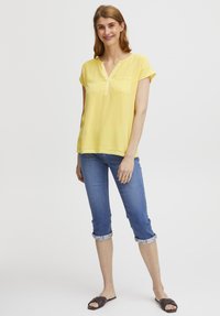 Gul kortærmet top med en klapnack og brystlomme, parret med blå jeans med opkrøllede ærmer og brune flade sandaler.