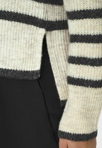Pull rayé en tricot doux. Présente des rayures beige clair et gris foncé, des poignets côtelés, des fentes latérales et une coupe décontractée.