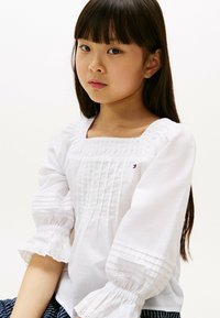 Blusa bianca di cotone con maniche a sbuffo, scollatura quadrata e dettagli plissettati. Presenta una delicata rifinitura ad occhio sulle estremità delle maniche.