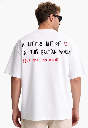 Homme portant un T-shirt blanc avec le texte "A little bit of love in this brutal world (but not too much)" au dos, avec un petit symbole de cœur rouge.