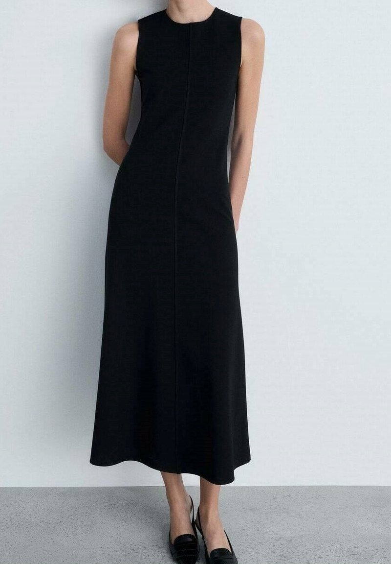 Robe midi noire sans manches avec encolure ronde, portée avec des escarpins noirs à bride arrière sur un sol gris contre un mur blanc.