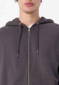 Mørkegrå hettegenser med glidelås, kengurulomme, snøring på hetten og sølvglidelås. Stoffet fremstår mykt med en jevn tekstur.