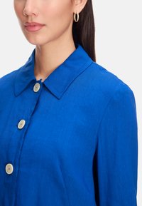 Blue linen shirt featuring a pointed collar, large cream buttons, and a smooth texture. Close-up of the neckline and fabric details.